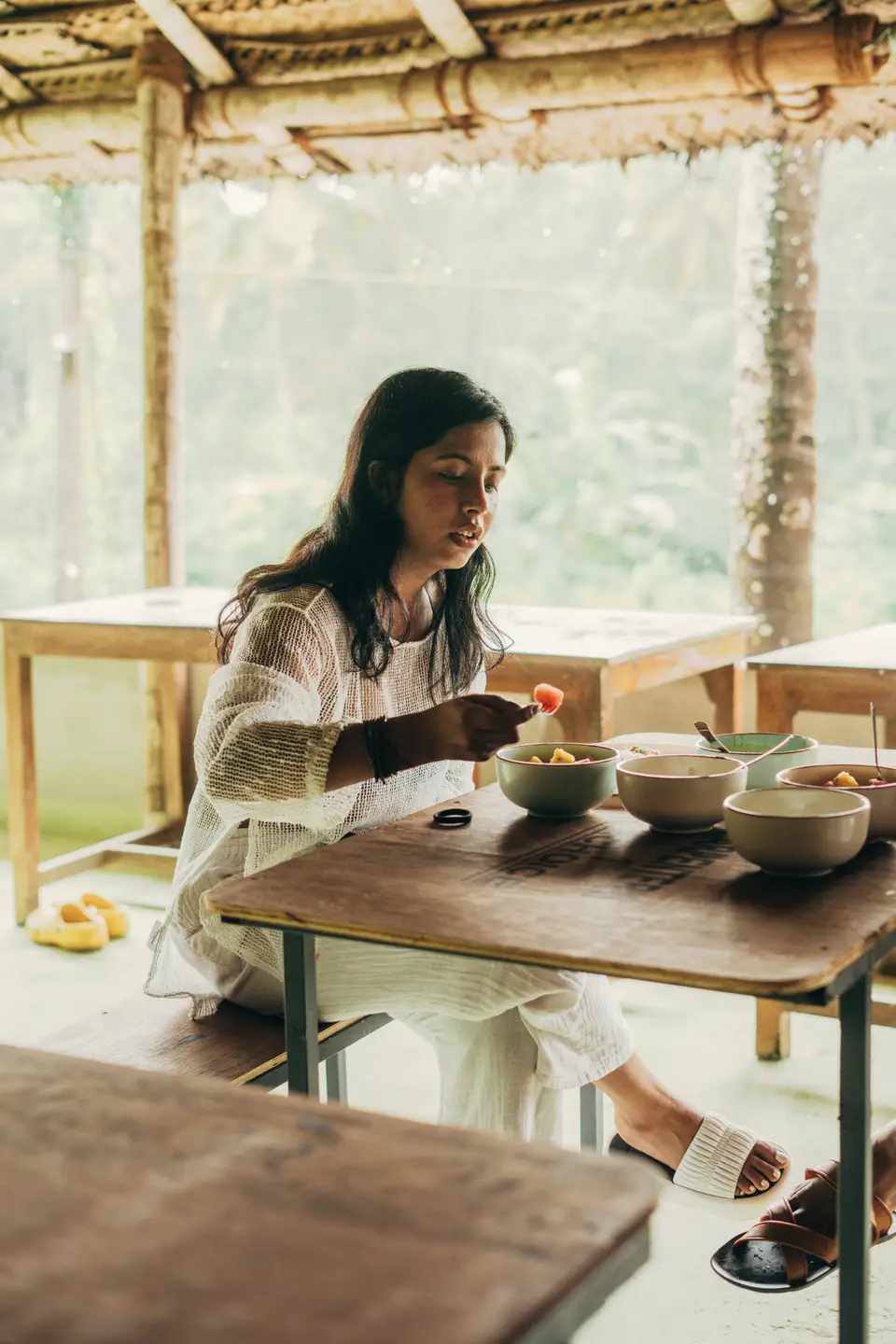 A woman in white eats cut fruit at a wooden table set with ceramic bowls, under a thatched roof with trees beyond
