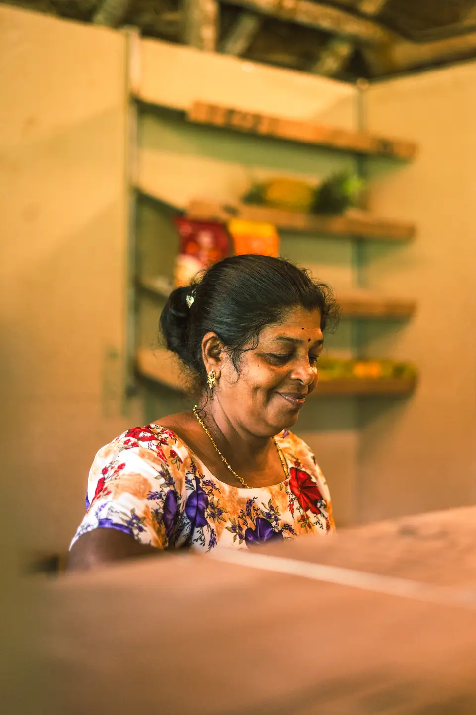 A cook smiling behind the counter of a small kitchen with shelves of jars