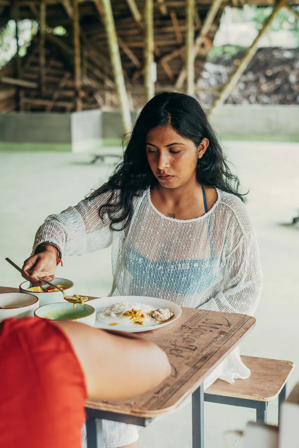A woman in a white knit top spoons curry from a bowl onto a plate of string hoppers at a wooden table