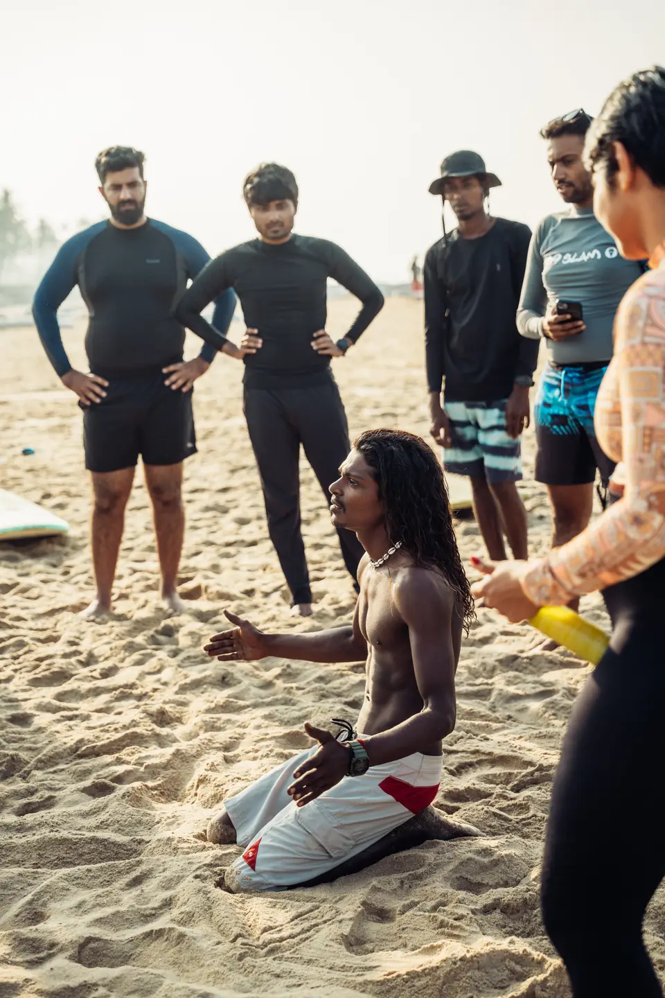 Kuma kneeling on the sand, giving a surf lesson to a group standing round him
