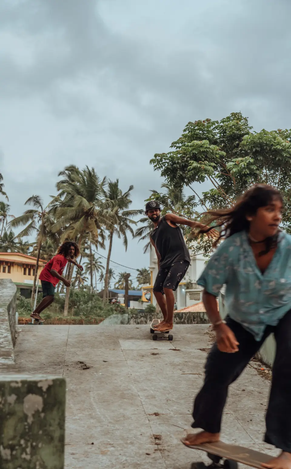 Three people skate barefoot down a concrete slope under a grey sky, with palms and houses behind