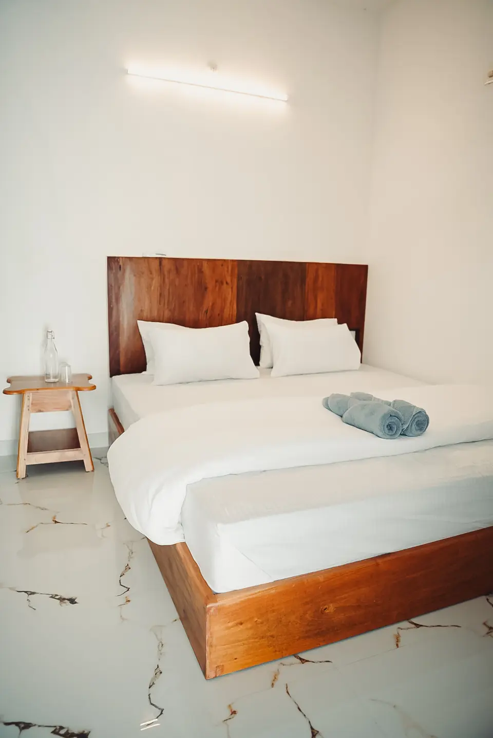 A bed with a wooden headboard, white pillows and rolled grey-blue towels beside a stool holding a glass water bottle