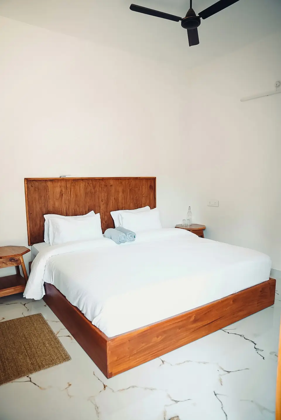A made-up double bed with a wooden headboard, white linen, rolled blue-grey towels and a jute mat on the floor