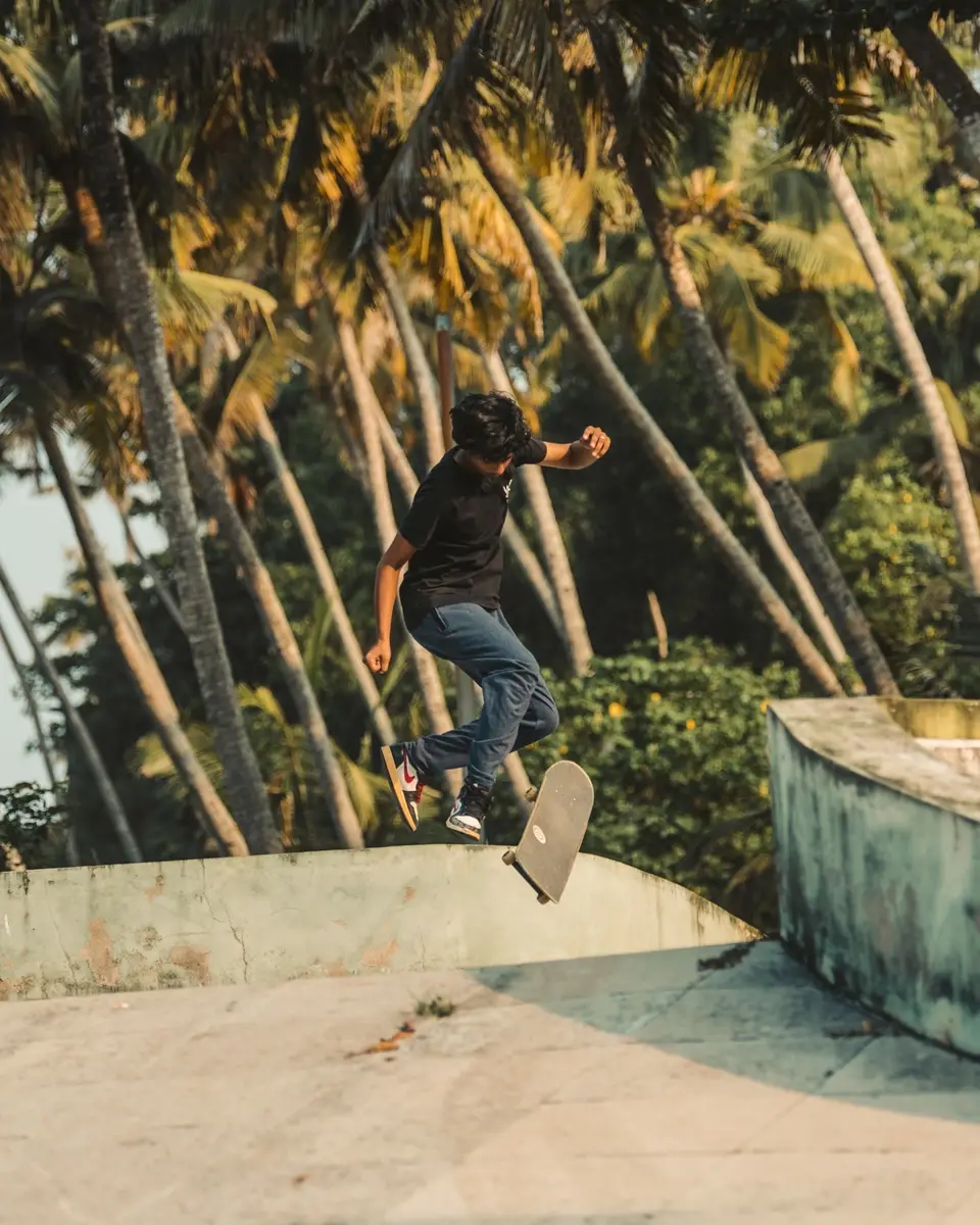A skater in a black tee and jeans mid-air above his flipping skateboard, a curved concrete wall and palms behind