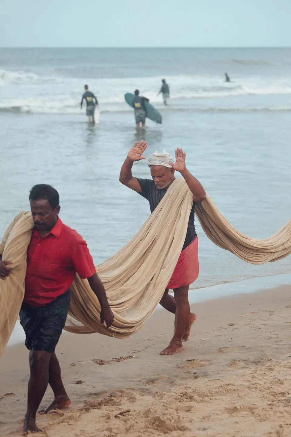 Two fishermen carrying a long beige fishing net along wet sand while surfers wade into the sea behind them