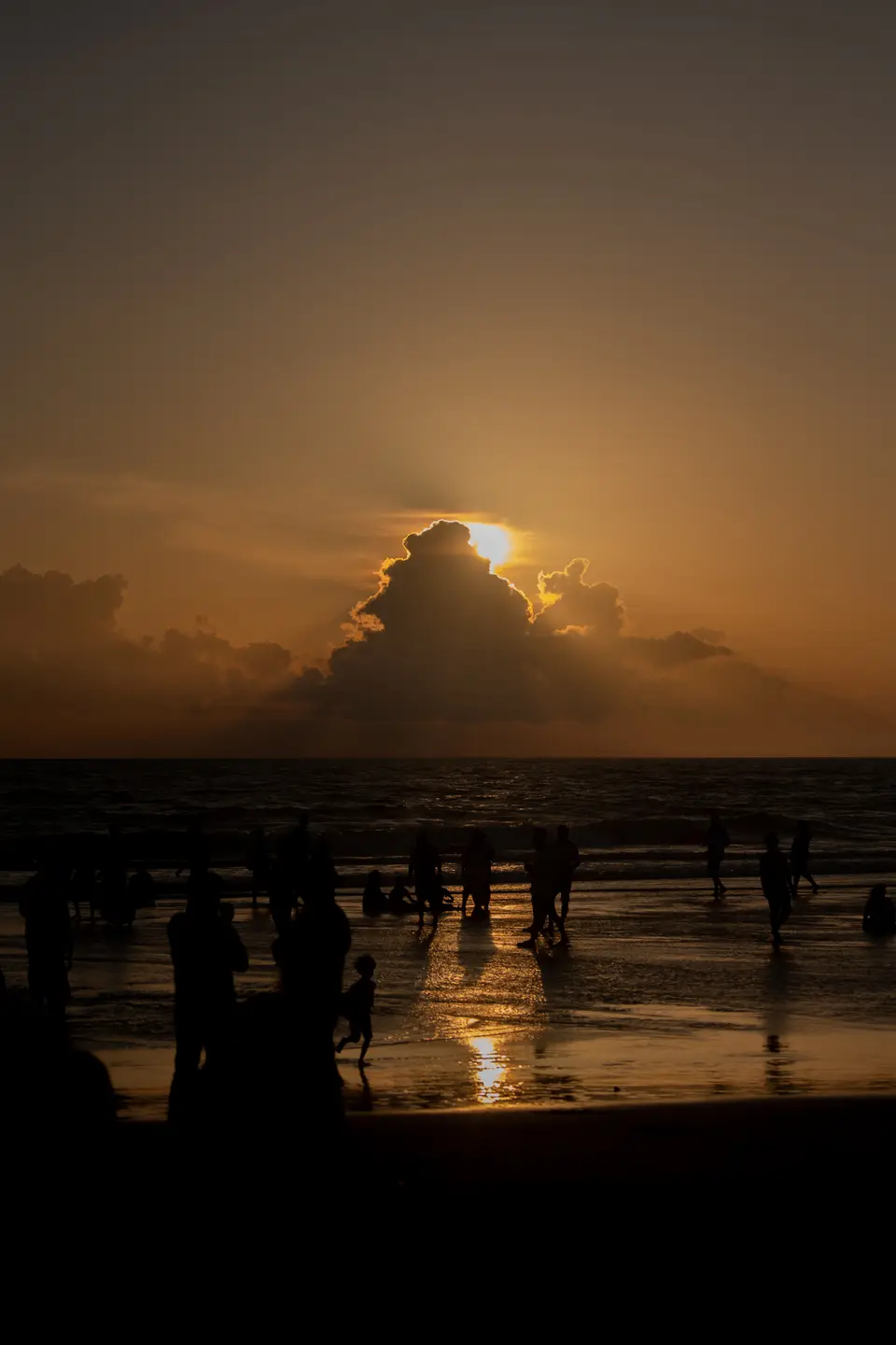 People silhouetted on the wet sand at sunset, the sun behind a cloud over the sea