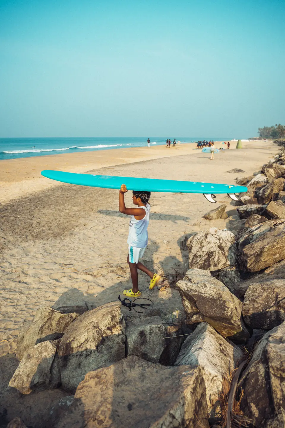 A child carrying a surfboard on their head across the rocks at the edge of the beach