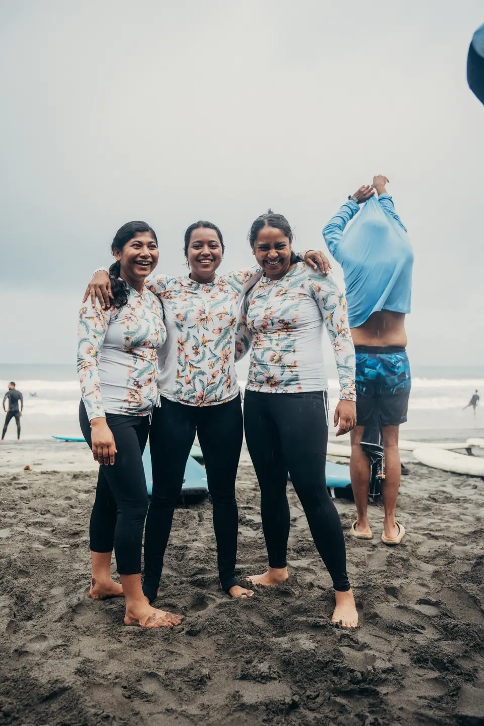 Three smiling women in matching floral rash guards stand arm in arm on dark sand, boards and grey sea behind