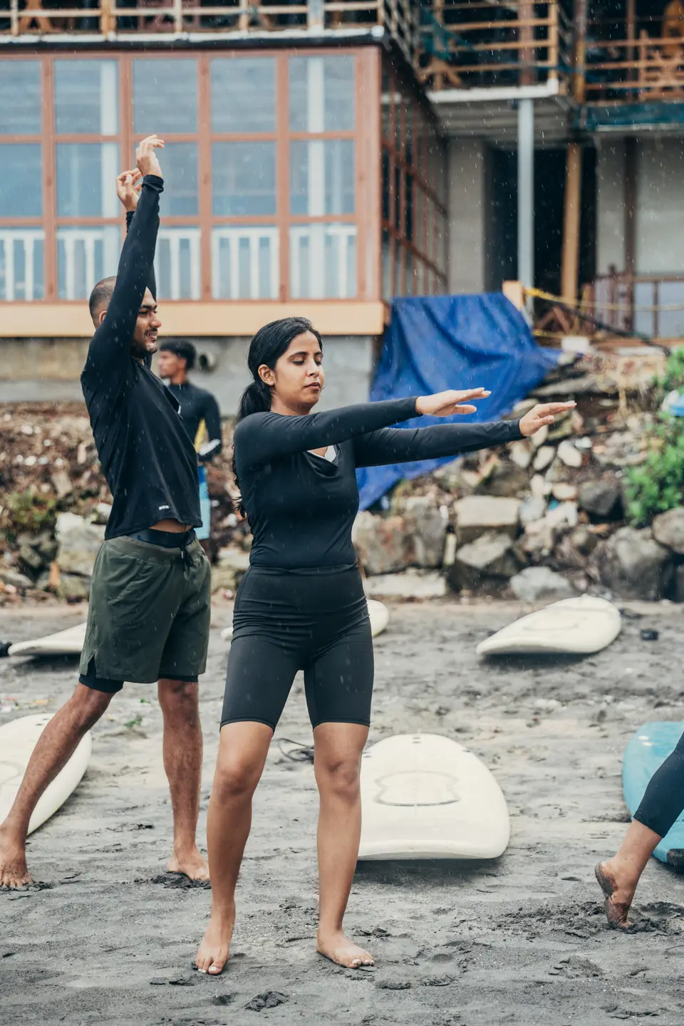 A man and a woman in black rash guards warm up on dark sand with arms raised, soft-top boards laid out around them