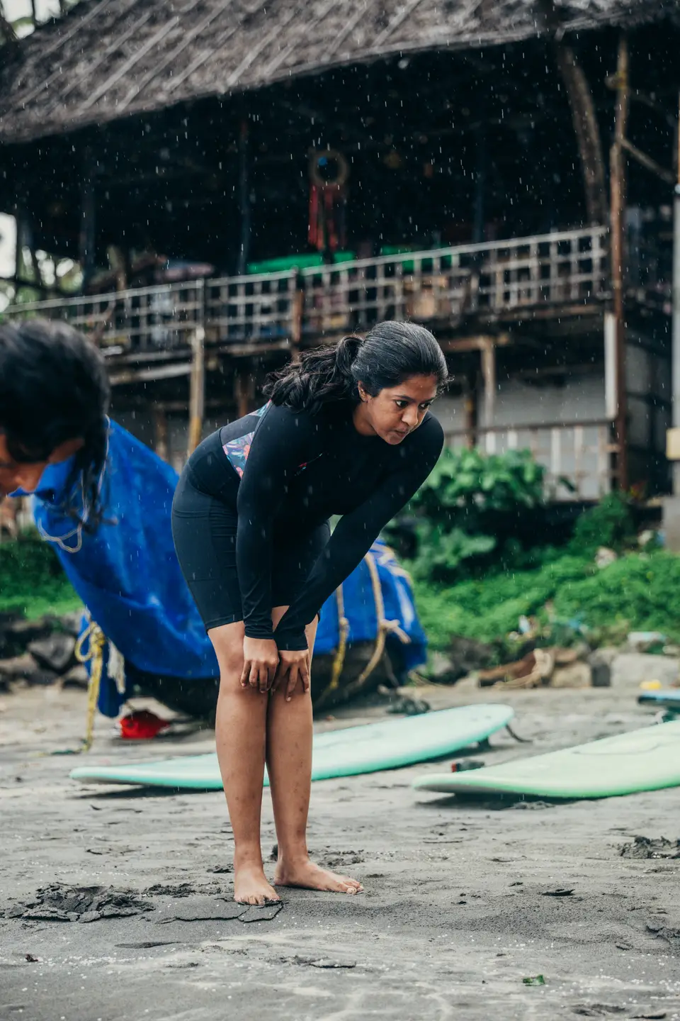 A woman in a black rash guard bends forward, hands on knees, beside mint-green soft-tops on dark sand in the rain