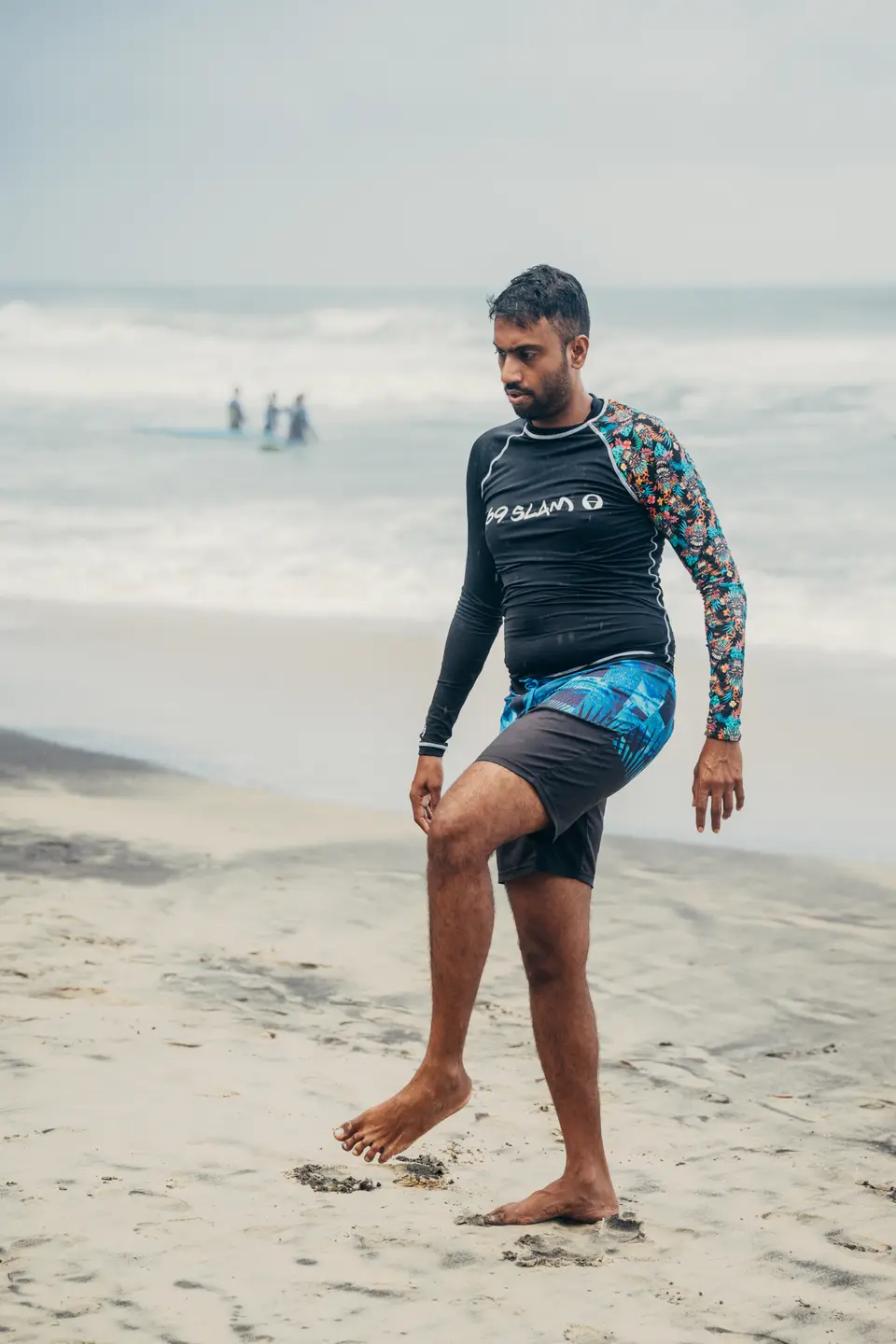 A man in a rash vest lifting one knee in a warm-up on the sand, the sea grey behind him