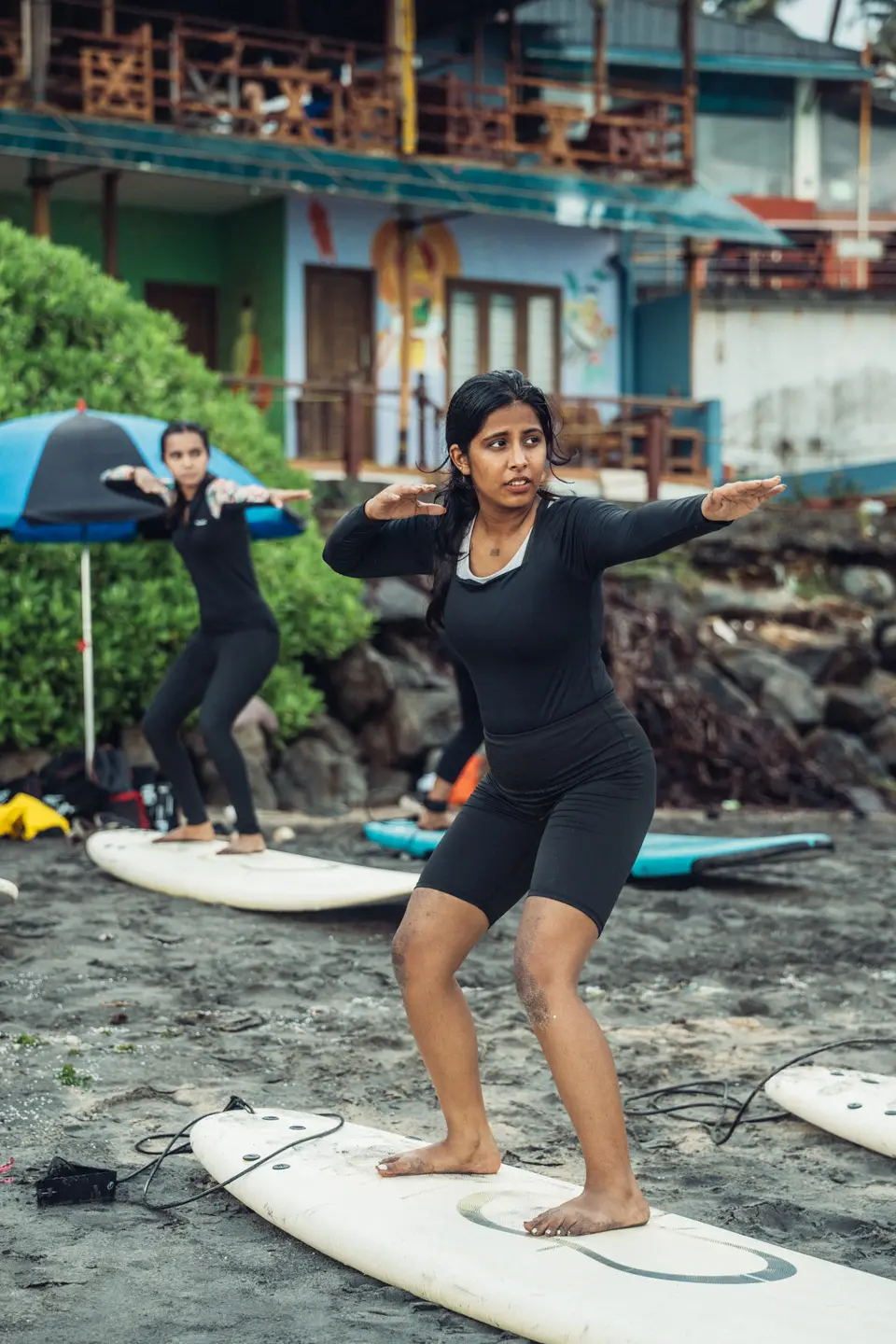A woman in black holds a pop-up stance on a soft-top laid on the sand while another woman behind holds the same pose