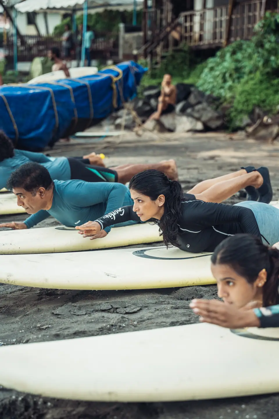 A row of people lying on white soft-top boards on the sand, practising the paddle position