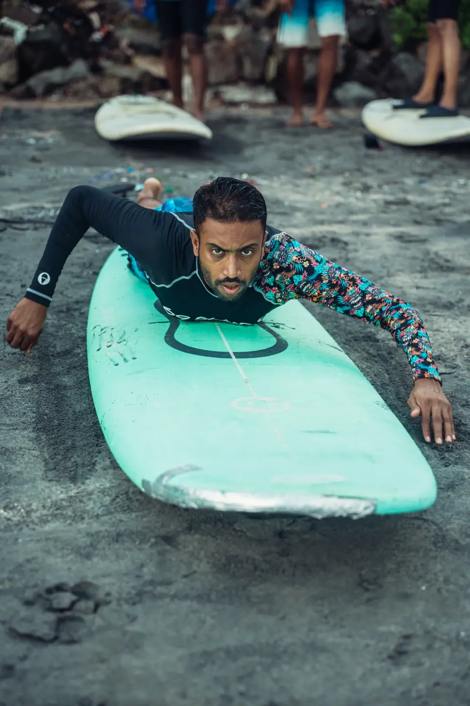 Close-up of someone lying on a mint soft-top board on dark sand, hands flat either side, the board's nose scuffed