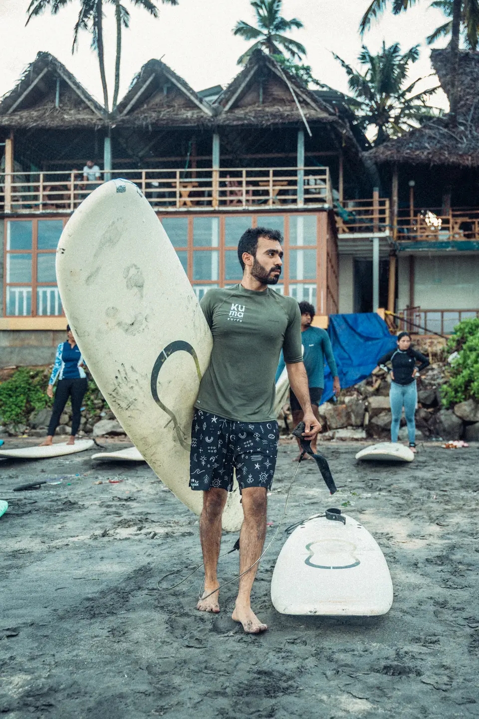 A bearded man in a Kuma rash guard holds a white board under one arm, leash in hand, another board at his feet