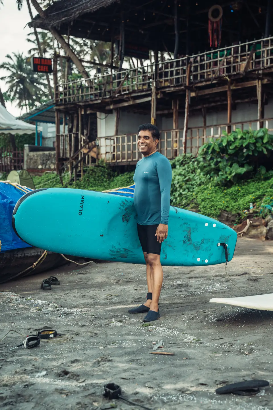 A smiling man in a teal rash guard, black shorts and neoprene booties holds a turquoise soft-top at his side on dark sand