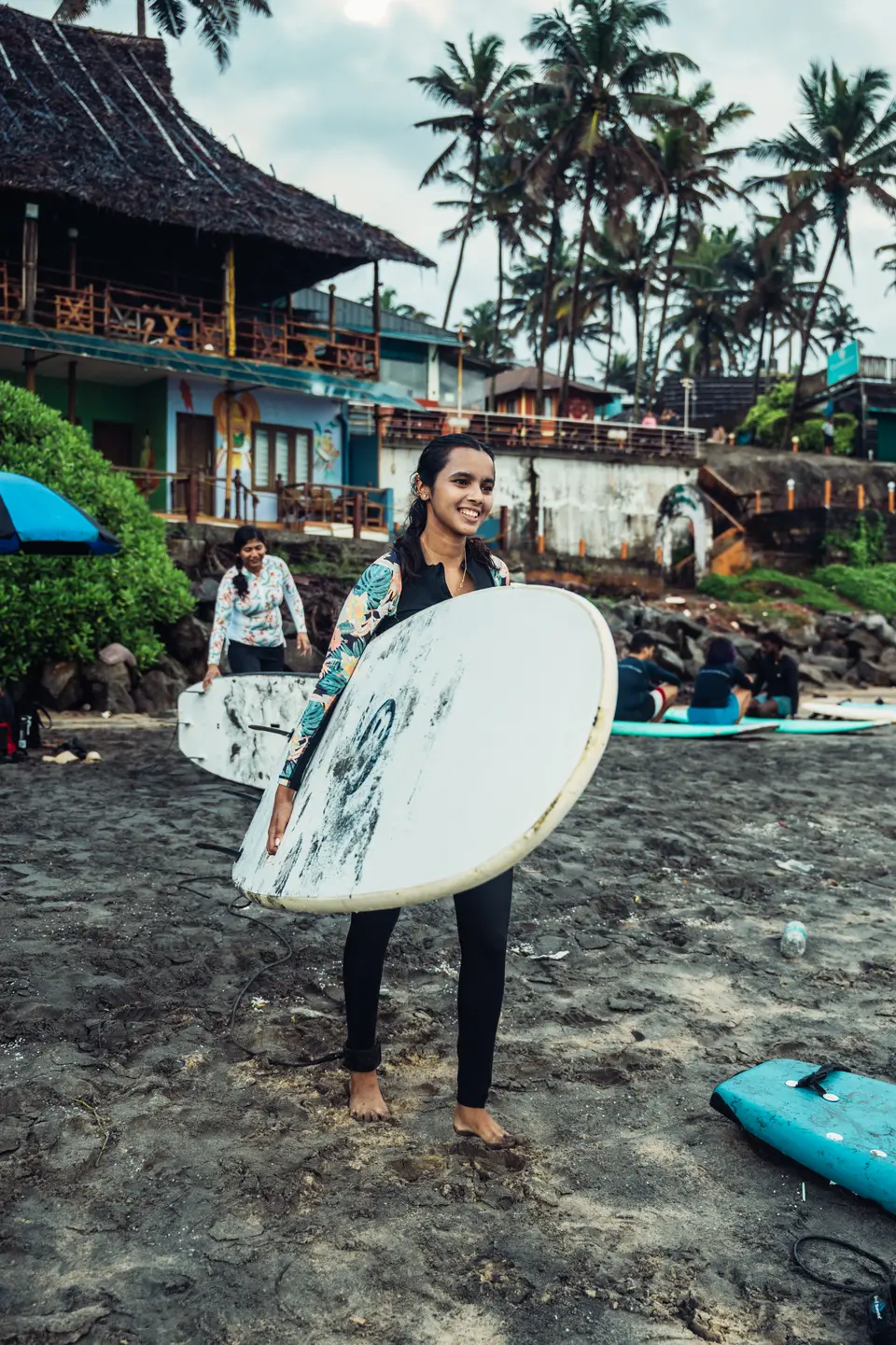 A smiling woman carries a white soft-top across dark sand, a thatched building, palms and people on boards behind her