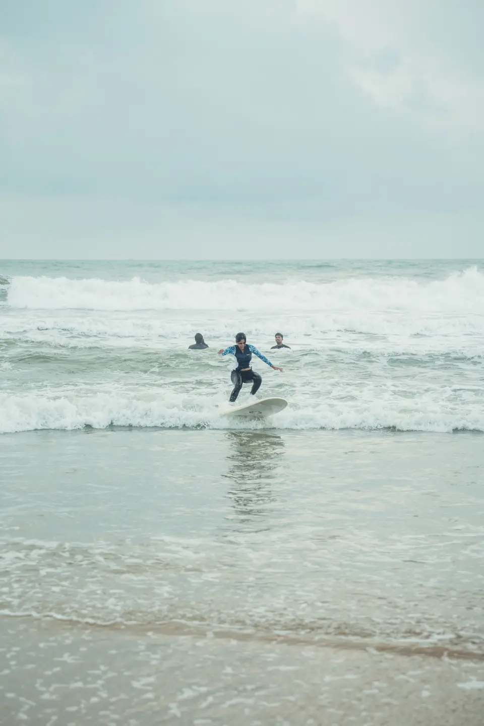 A distant surfer rides whitewater on a white board across a grey sea, two heads in the water behind