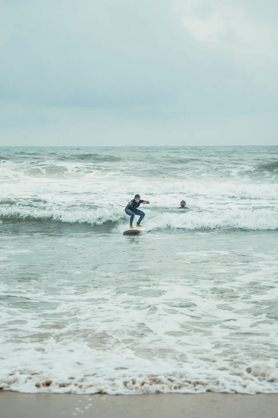 A distant surfer in dark long sleeves crouches on a board in broken whitewash, one other person in the water nearby