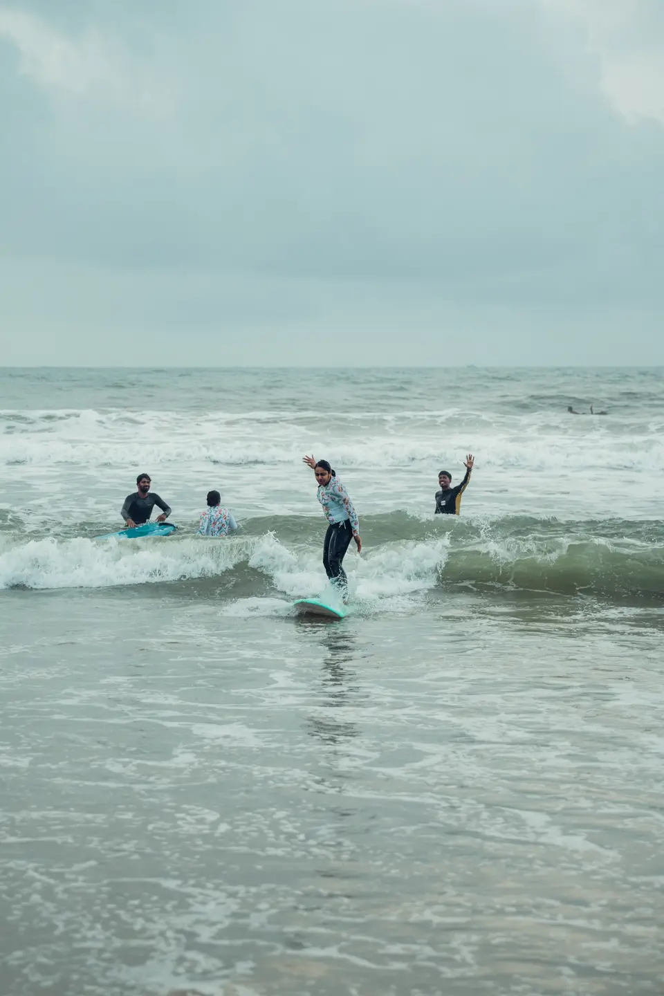 A woman rides whitewater on a teal soft-top with three people in the water behind her, one of them waving