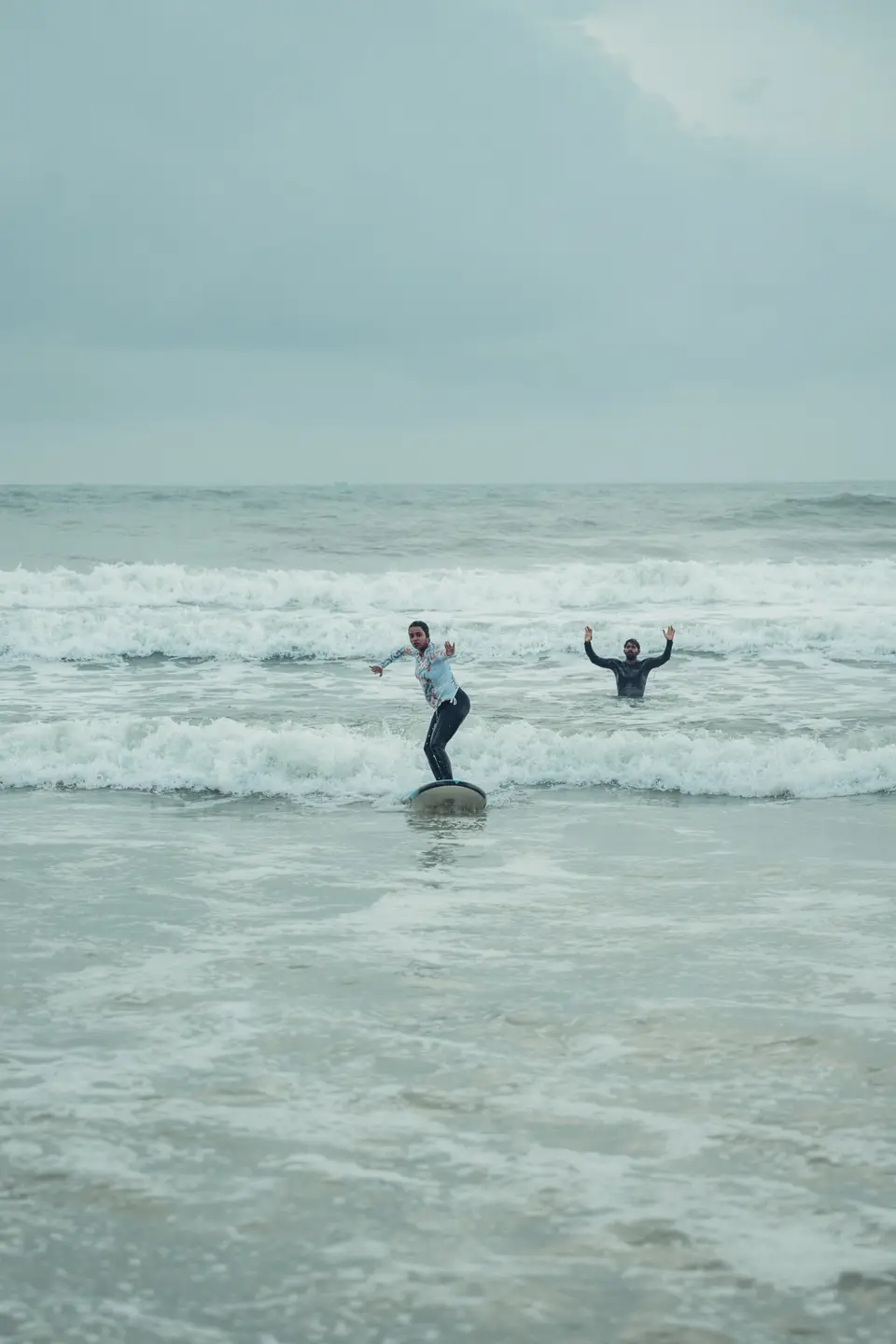 A woman in a light rash guard stands on a board in whitewash while a man behind her raises both arms