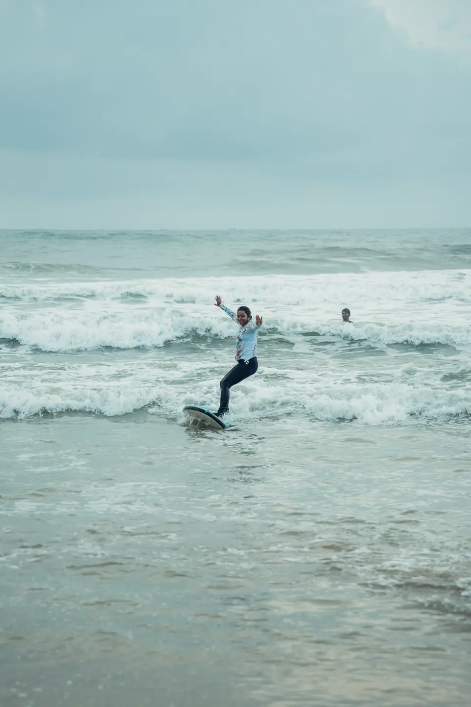 A woman in a floral rash guard rides whitewater on a dark soft-top with both arms flung up, one head in the water behind