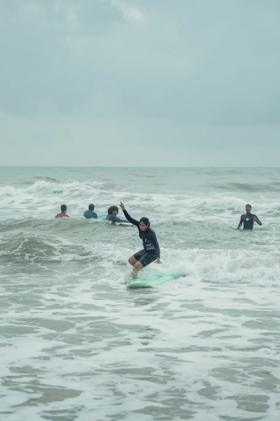 A woman in a black rash guard rides whitewater on a mint-green soft-top while others wait in the water behind her