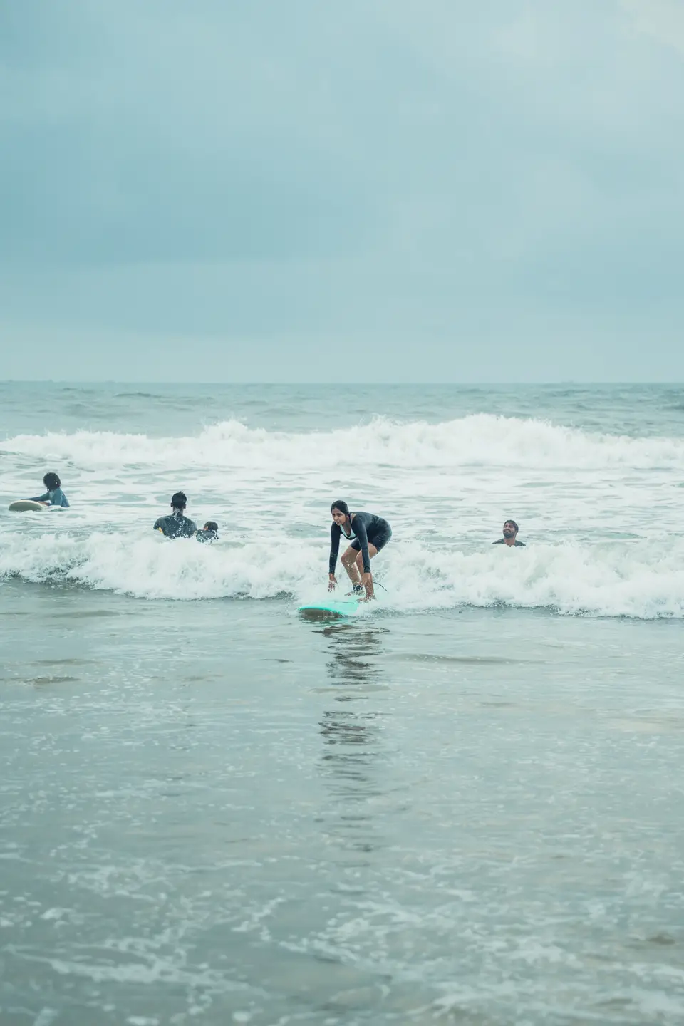 A woman in a black rash guard rides whitewash on a teal soft-top toward the camera, other people in the water nearby