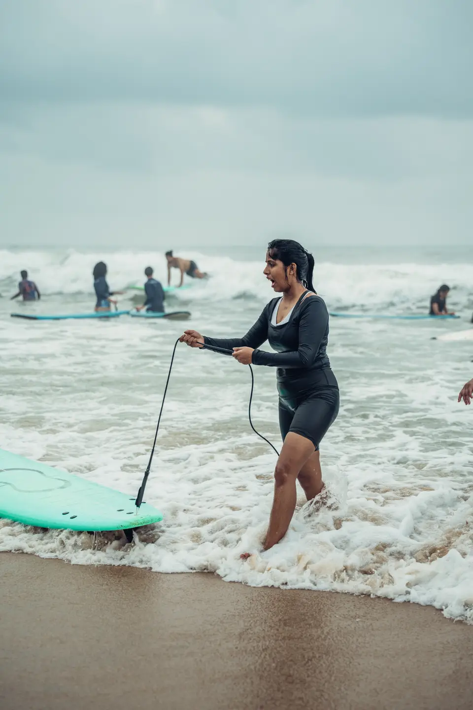 A woman in a black rash guard tows a mint soft-top through the shore break by its leash, mouth open