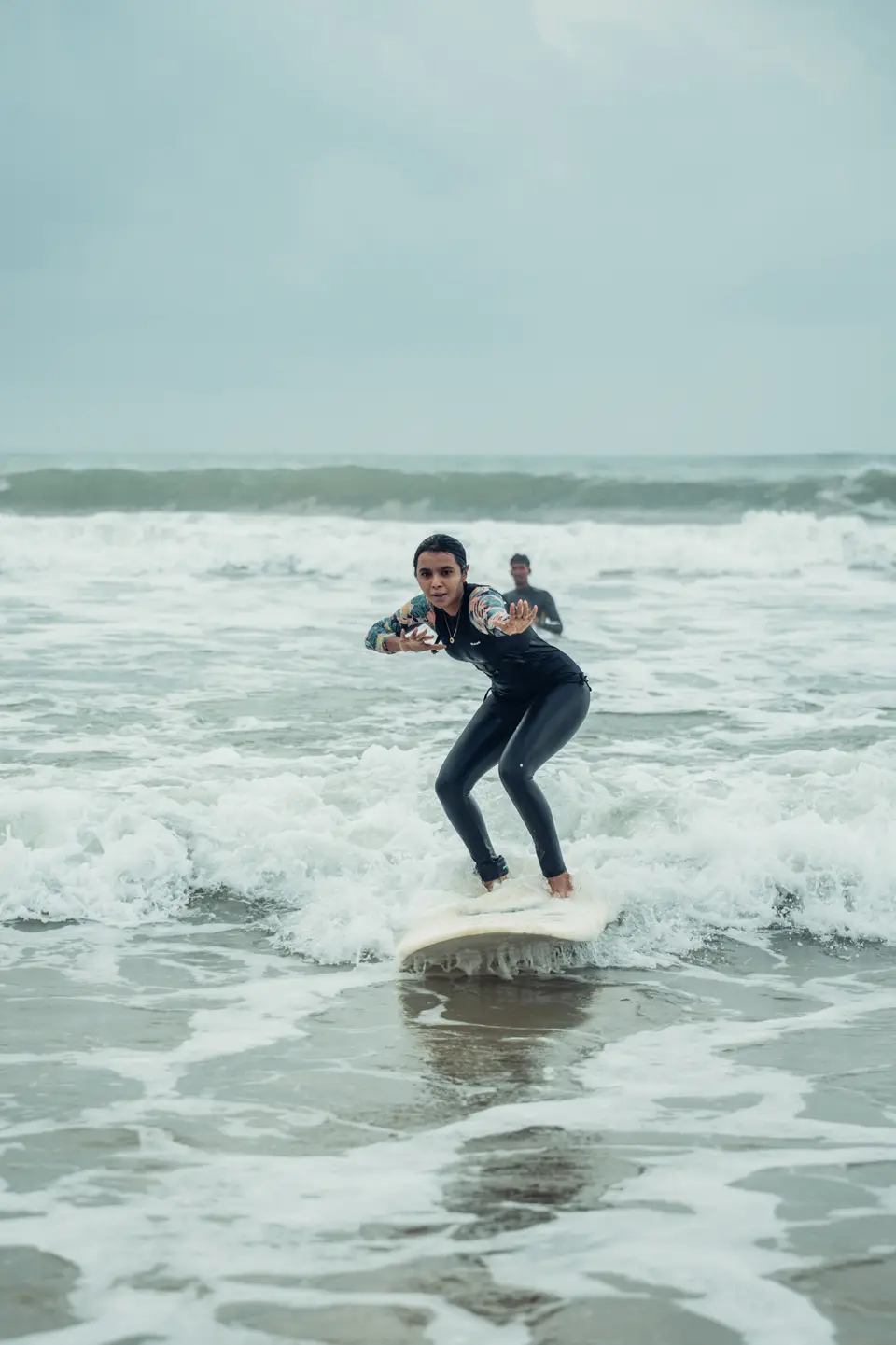 A woman in a floral-sleeved wetsuit stands up on a white board in the whitewater, a man waist-deep in the water behind her