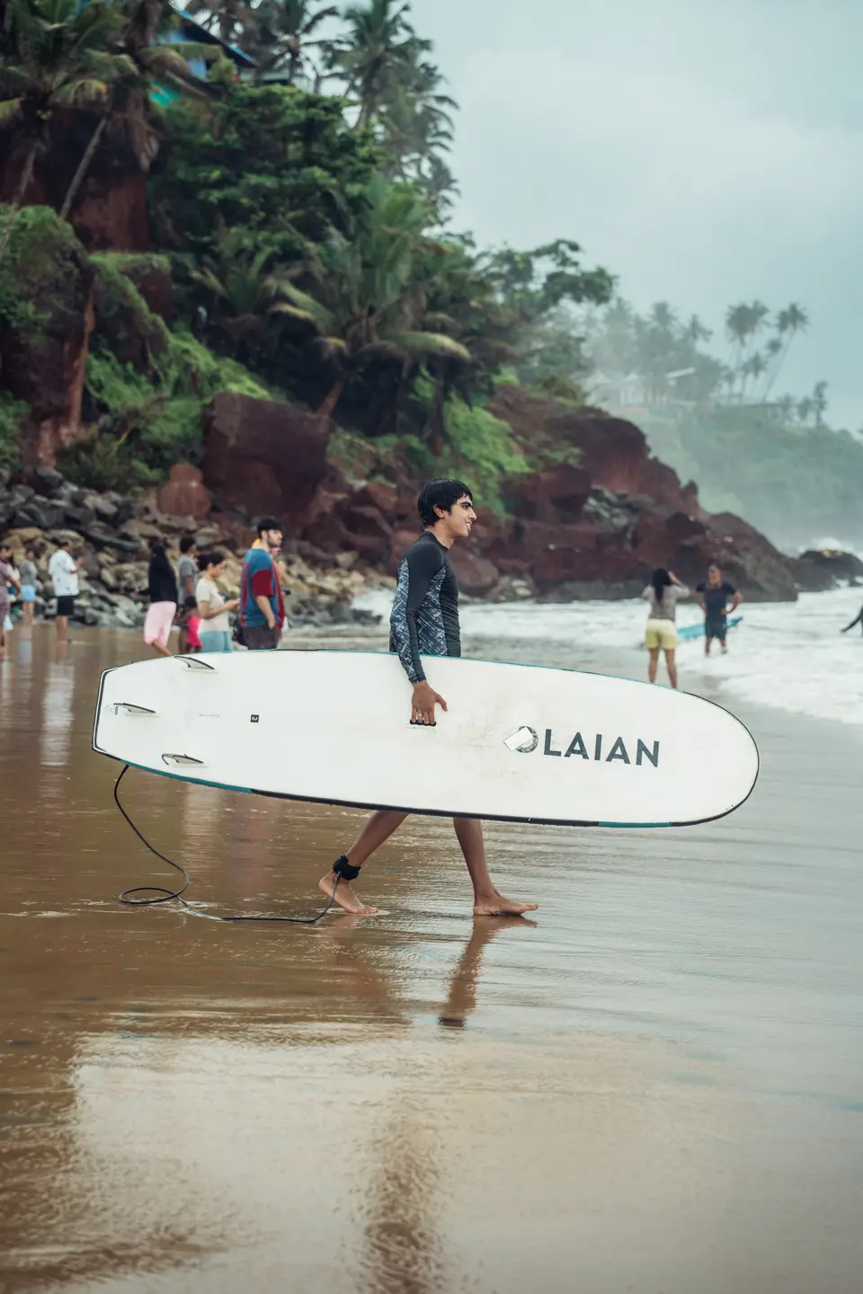 A man carries a large white board across wet sand, a crowd and a palm-covered red laterite cliff behind him