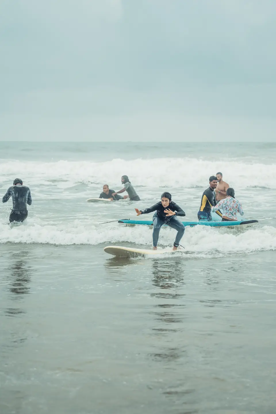 A woman in a black rash guard crouches on a cream board in shallow whitewash, a group with boards in the water behind