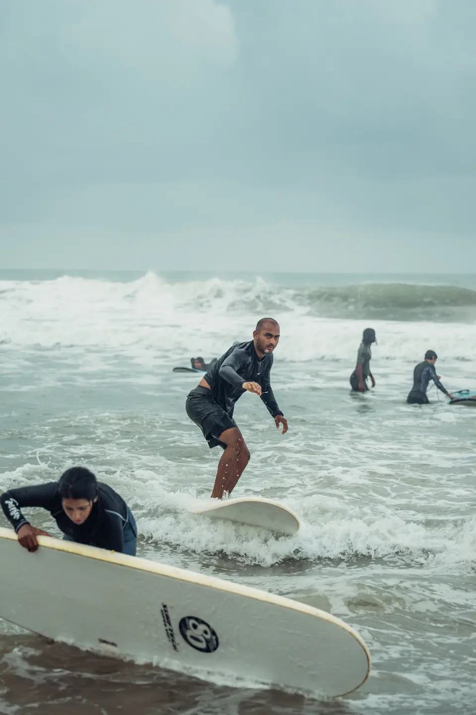 A man rides foam on a white soft-top as a woman in the foreground hauls another board out of the wash