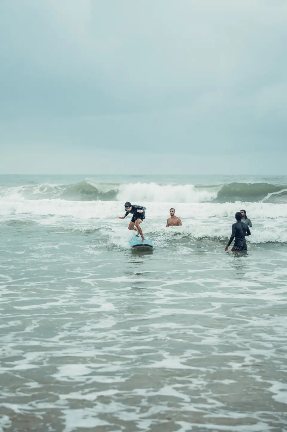 A surfer on a blue board rides whitewash as bigger waves break behind and people stand in the sea watching