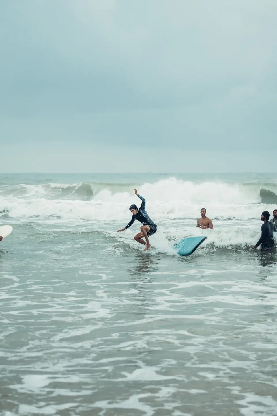 A surfer loses balance in whitewater, one arm up as the blue board kicks out, with three men in the water watching