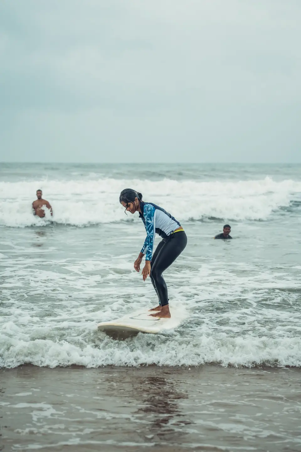 A woman in a palm-print rash guard stands on a cream board in whitewash, knees bent, looking at her feet