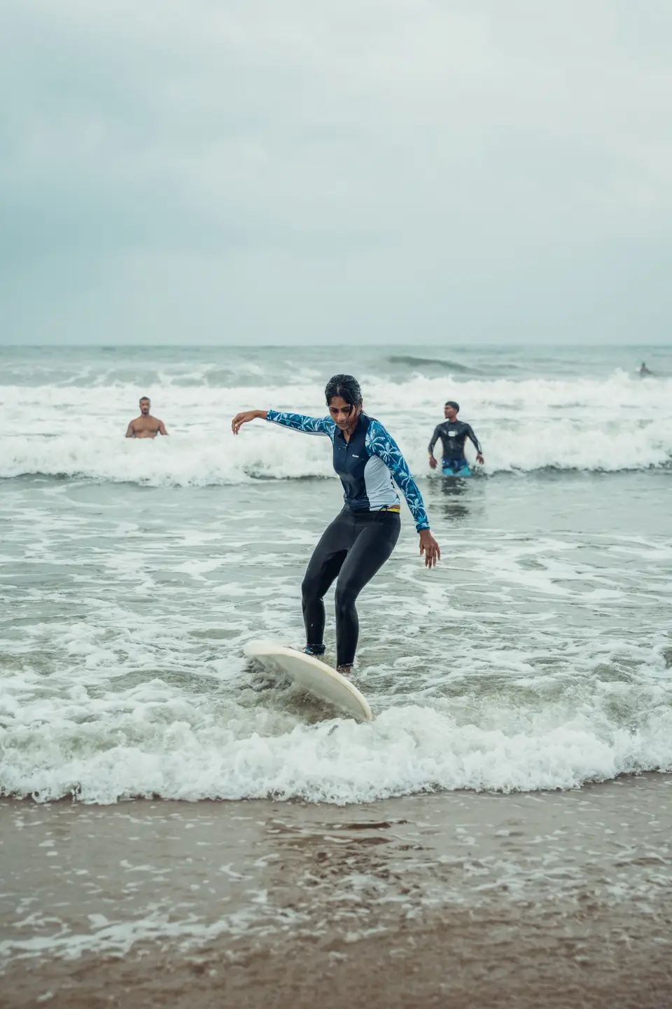 A woman in a floral-sleeved rash guard stands on a board in whitewash, arms out, two men in the water behind