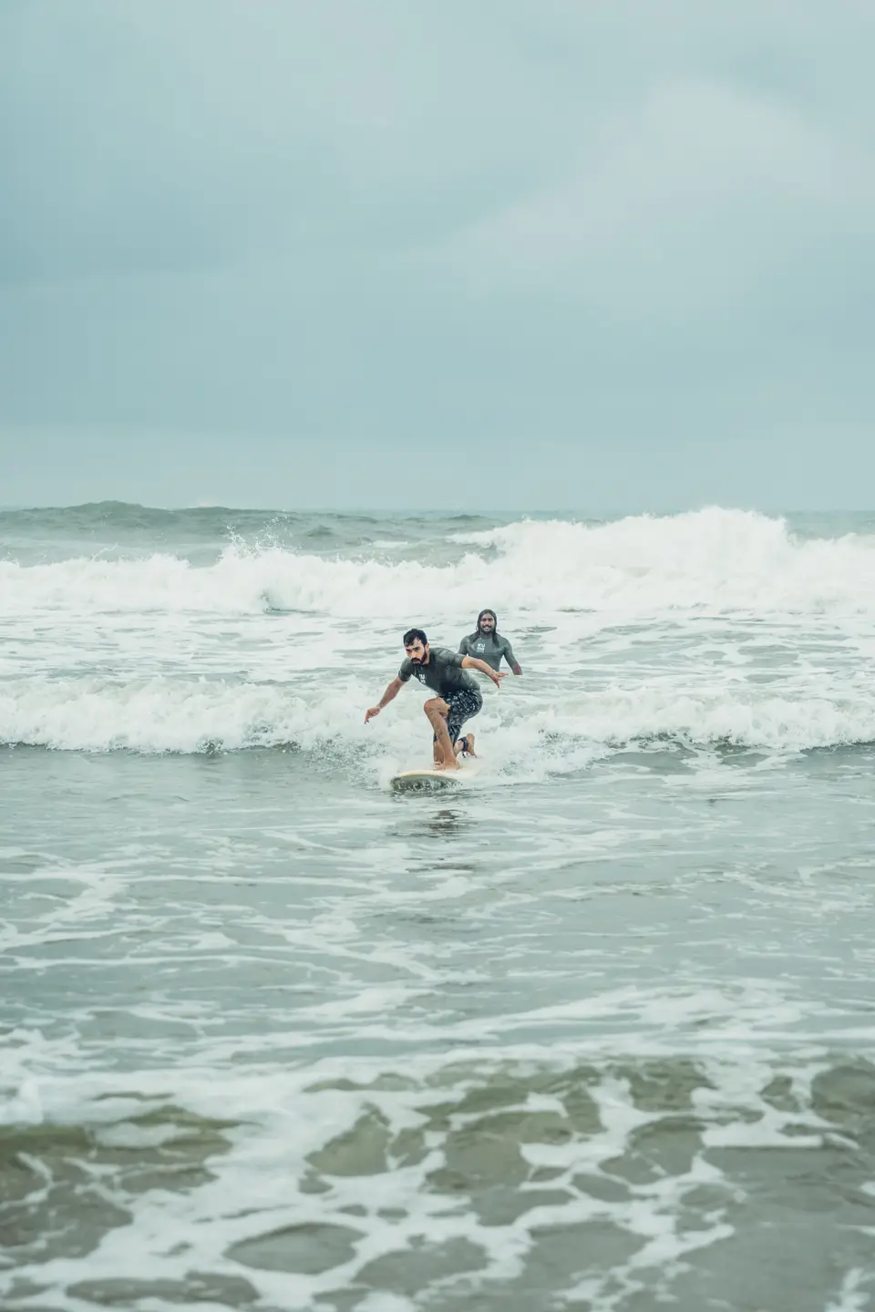 A bearded man crouches on a cream soft-top in whitewater, arms out, while a man in a grey rash guard stands behind