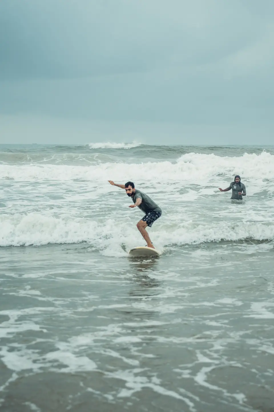 A bearded man in a grey rash guard and patterned shorts crouches on a board in whitewash, arms out for balance