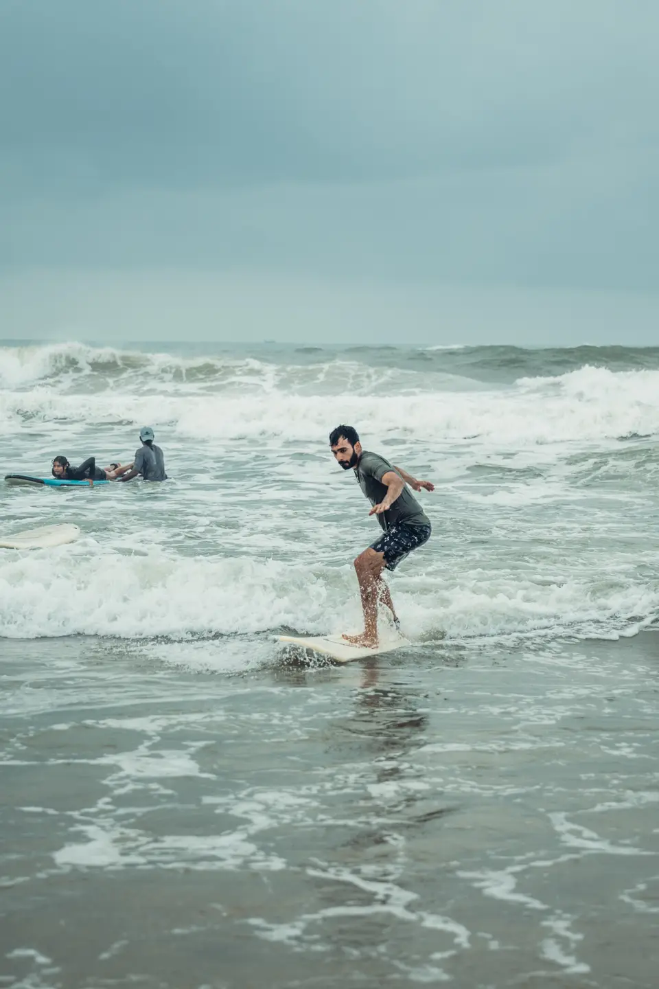 A bearded man stands on a white board in shallow whitewash with arms out, another surfer lying on a board behind