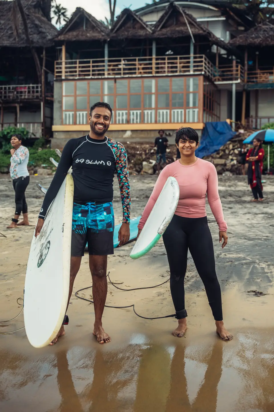 A smiling man and woman in rash guards hold white surfboards on wet sand, with a thatched multi-storey building behind