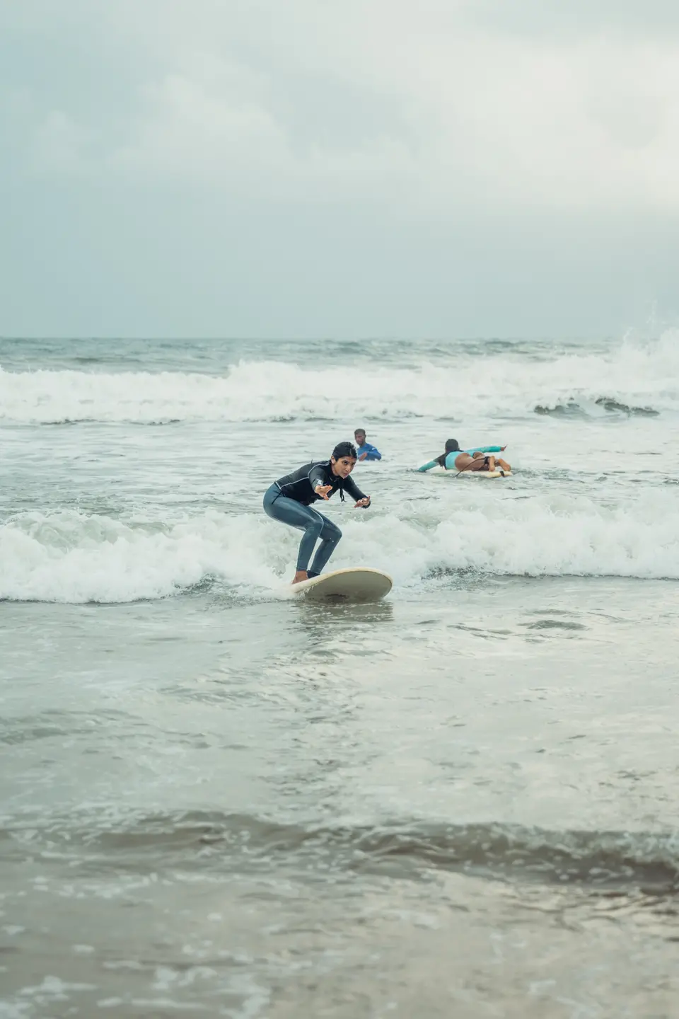 A woman in a black rash guard crouches low on a cream board in whitewash, others in the water behind her