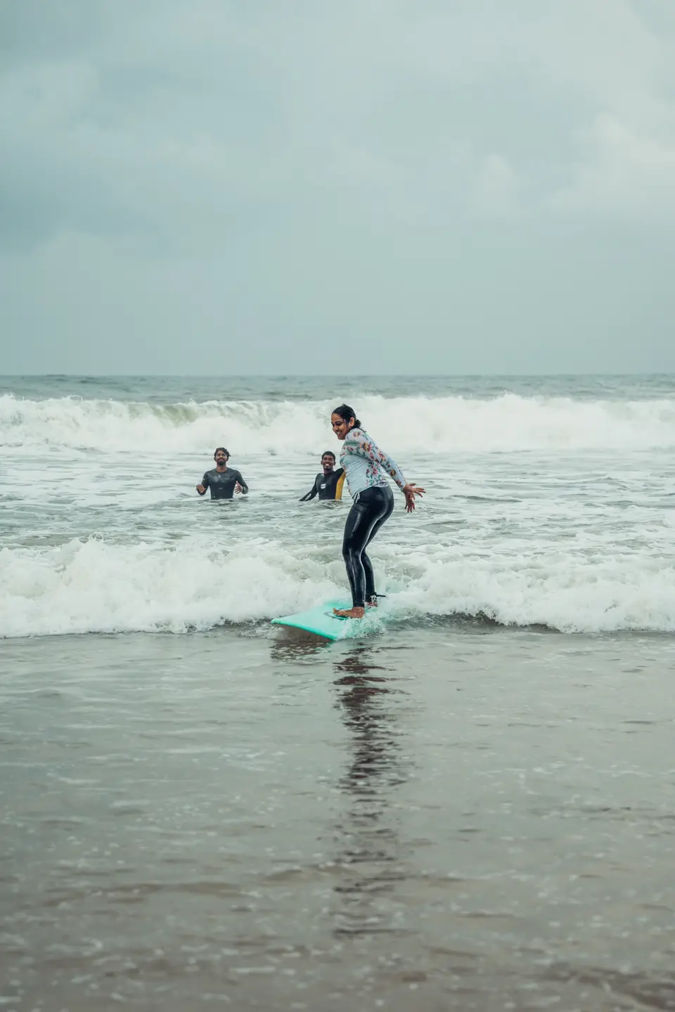 Up on a teal soft-top in the whitewash, two people in wetsuits watching from behind