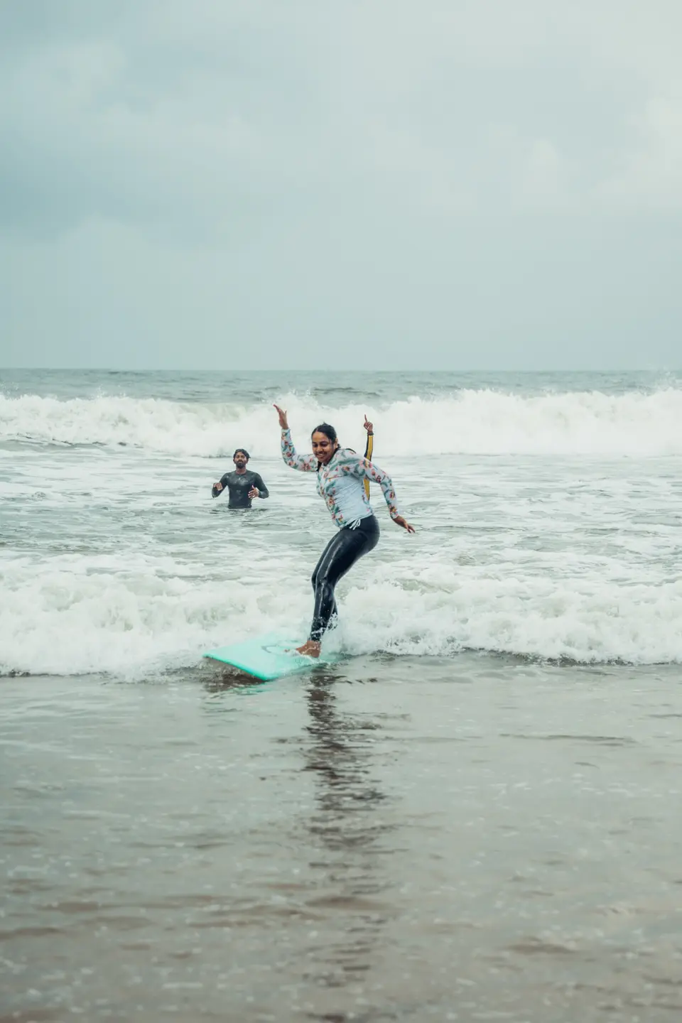 A smiling woman stands on a teal board in shallow whitewash, one arm up and pointing, a man in the water behind