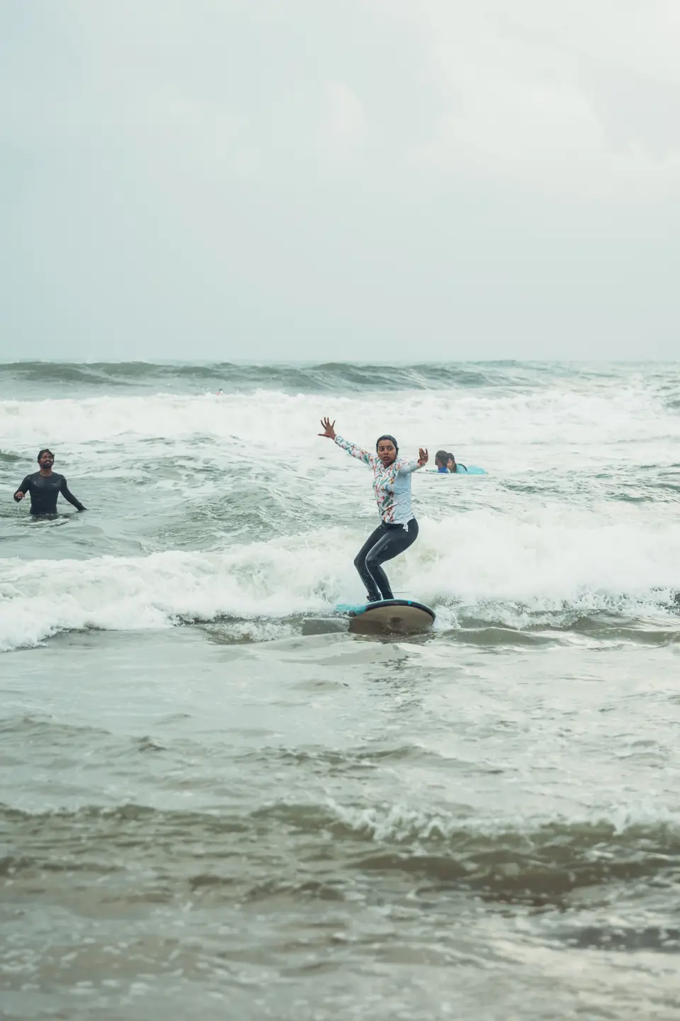 A woman in a white floral top stands on a board in choppy whitewash, both arms flung wide, a man in the water behind