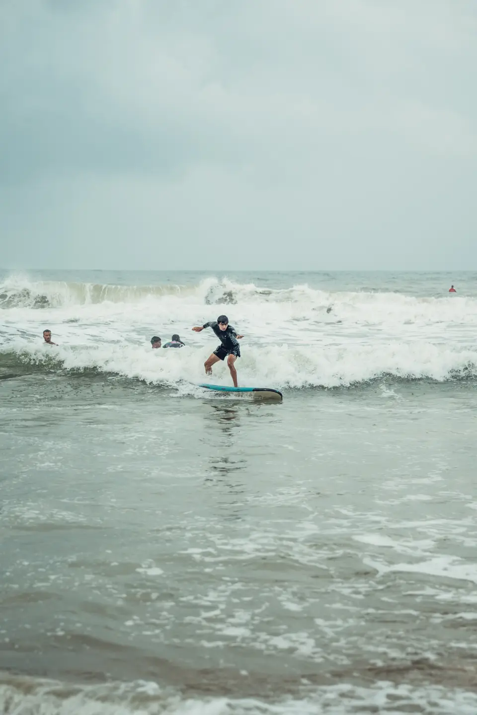 A surfer in a black rash guard rides whitewater on a blue soft-top, a few heads bobbing in the water nearby