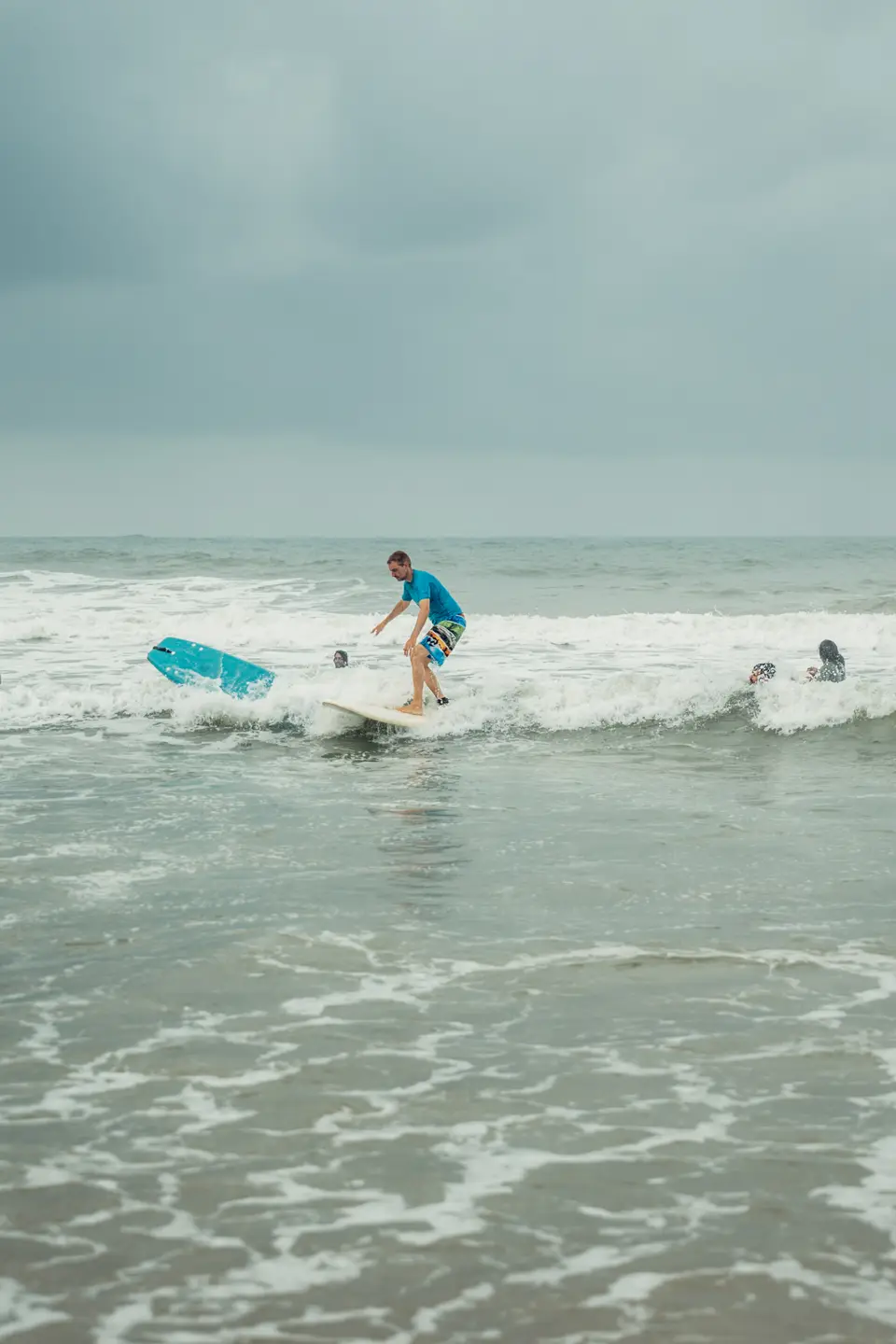 A man in a blue rash guard rides a white board through whitewash while a blue board flips through the air beside him