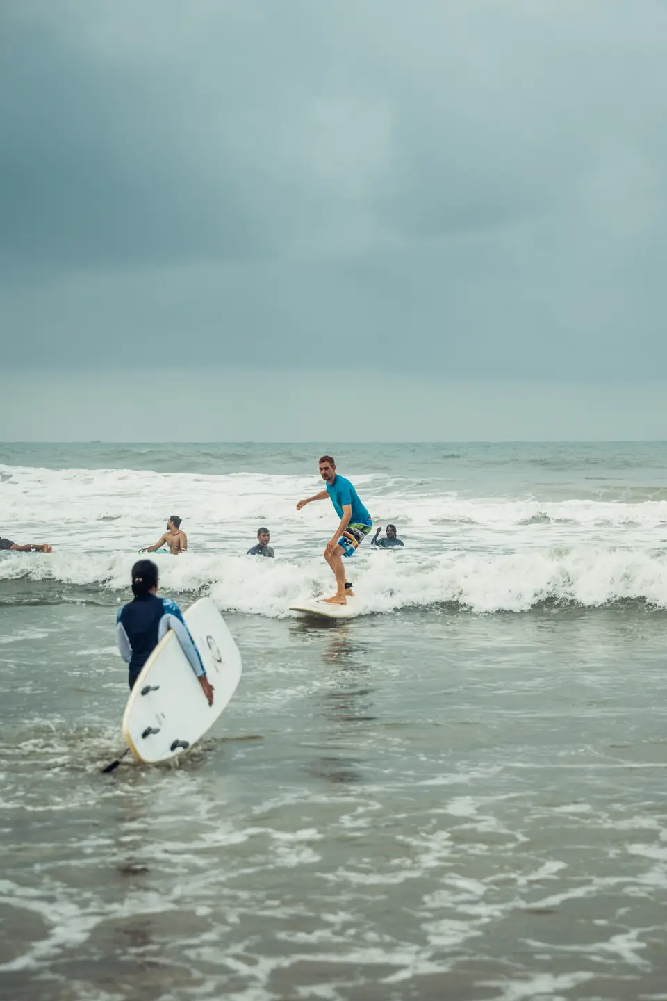 A man in a blue rash guard rides whitewater on a white soft-top while someone in the foreground wades out with a board