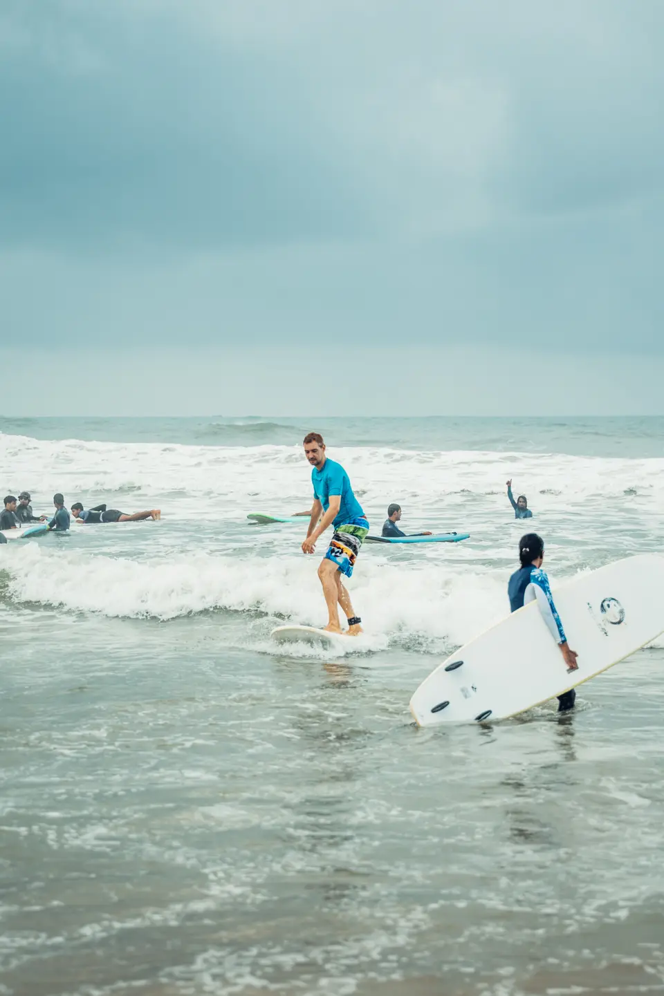 A man in a turquoise rash guard rides whitewash on a white board as someone carries another board past