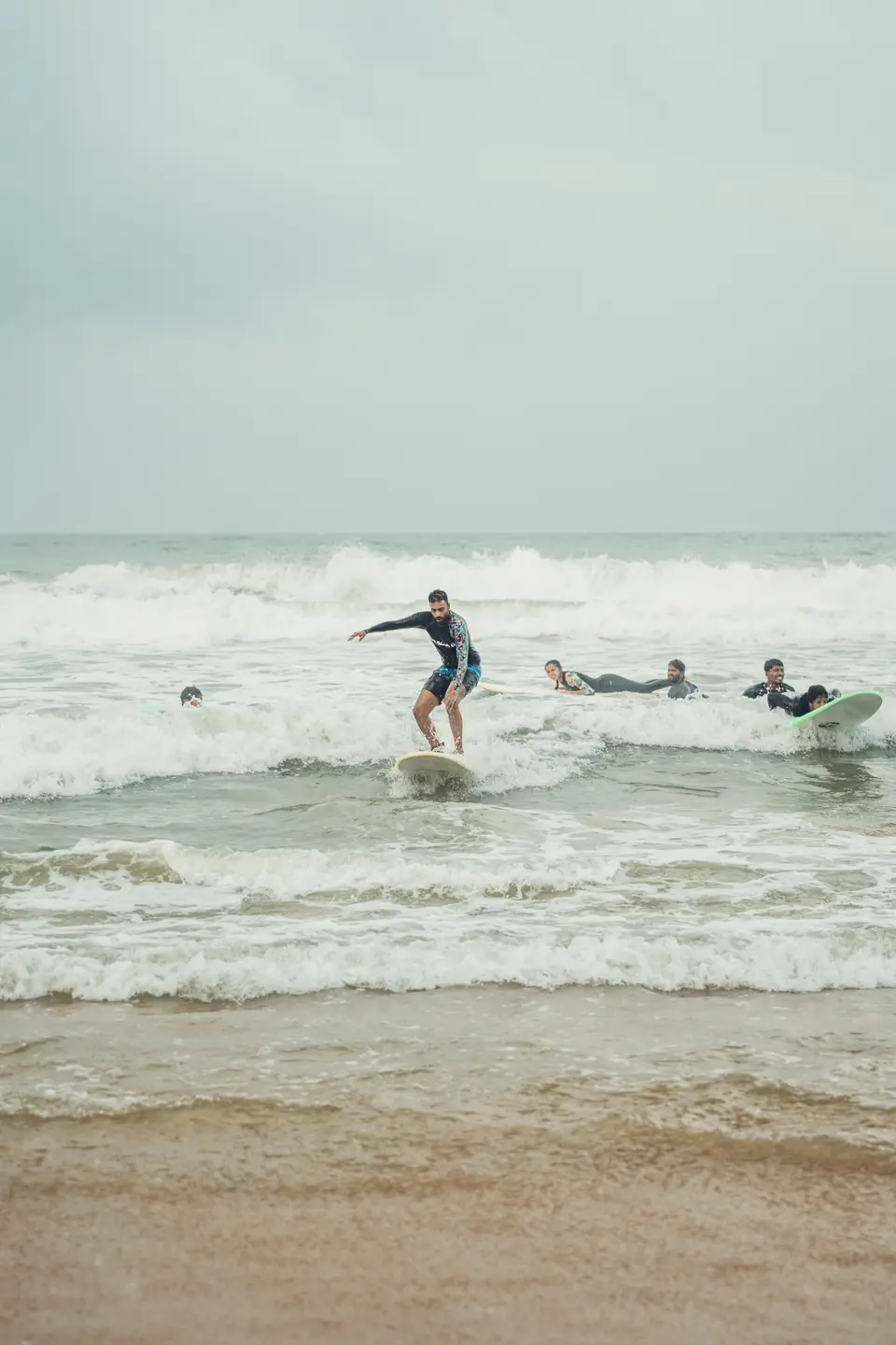 A man in a dark rash guard with patterned sleeves rides whitewater on a white board while others paddle out behind him